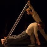 two acrobats both with brown hair and beards, wearing brown trousers and grey sweatshirts perform over a gym ball and with a cyr wheel in an indoor stage setting with yellow lighting