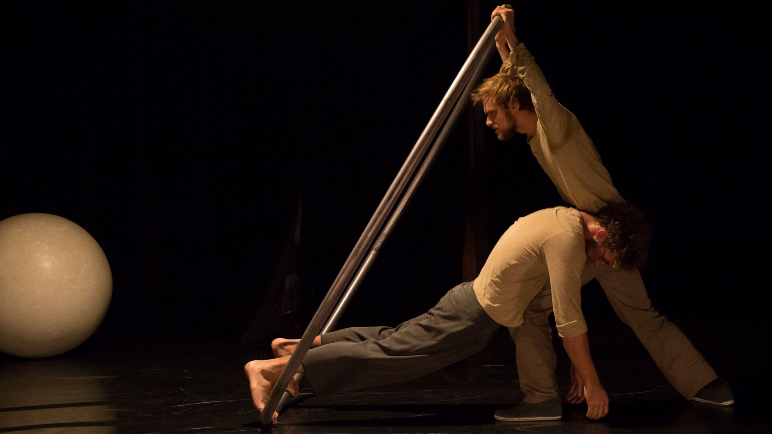 two acrobats both with brown hair and beards, wearing brown trousers and grey sweatshirts perform over a gym ball and with a cyr wheel in an indoor stage setting with yellow lighting