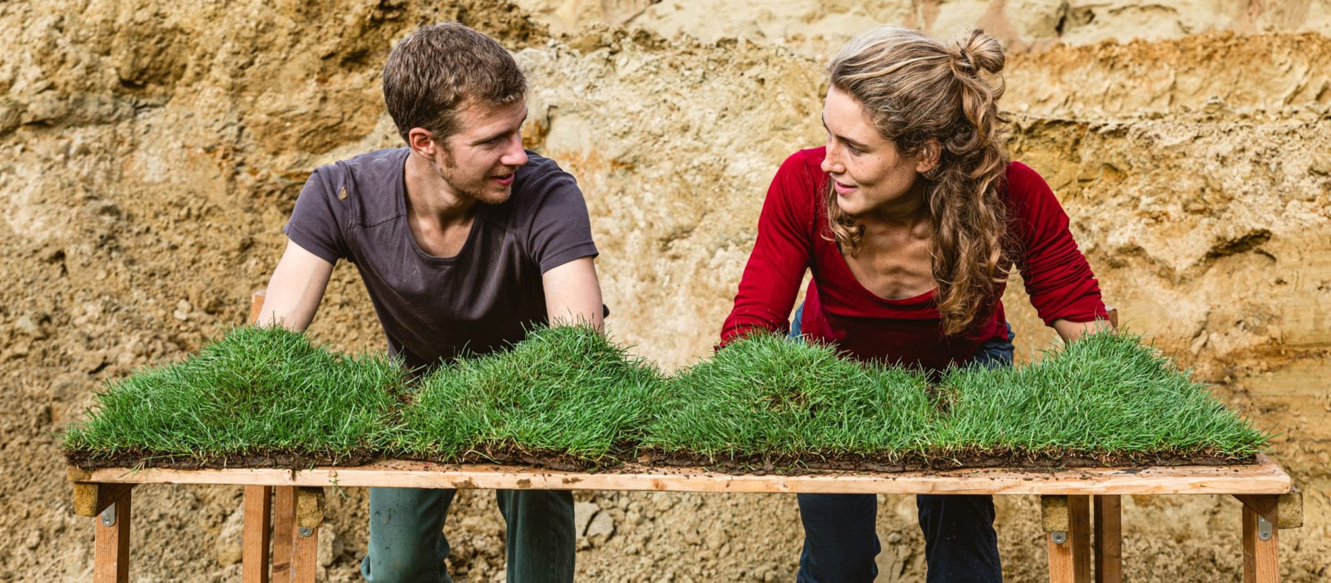 two performers in front of an outdoor muddy backdrop, one with long hair tied back and a red t-shirt, the other with short brown hair and a brown t-shirt, stand at a table with squares of grassy turf on it, they look at each other and have their arms and hands in the bright green turf summer 2023 recap