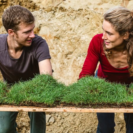 two performers in front of an outdoor muddy backdrop, one with long hair tied back and a red t-shirt, the other with short brown hair and a brown t-shirt, stand at a table with squares of grassy turf on it, they look at each other and have their arms and hands in the bright green turf summer 2023 recap