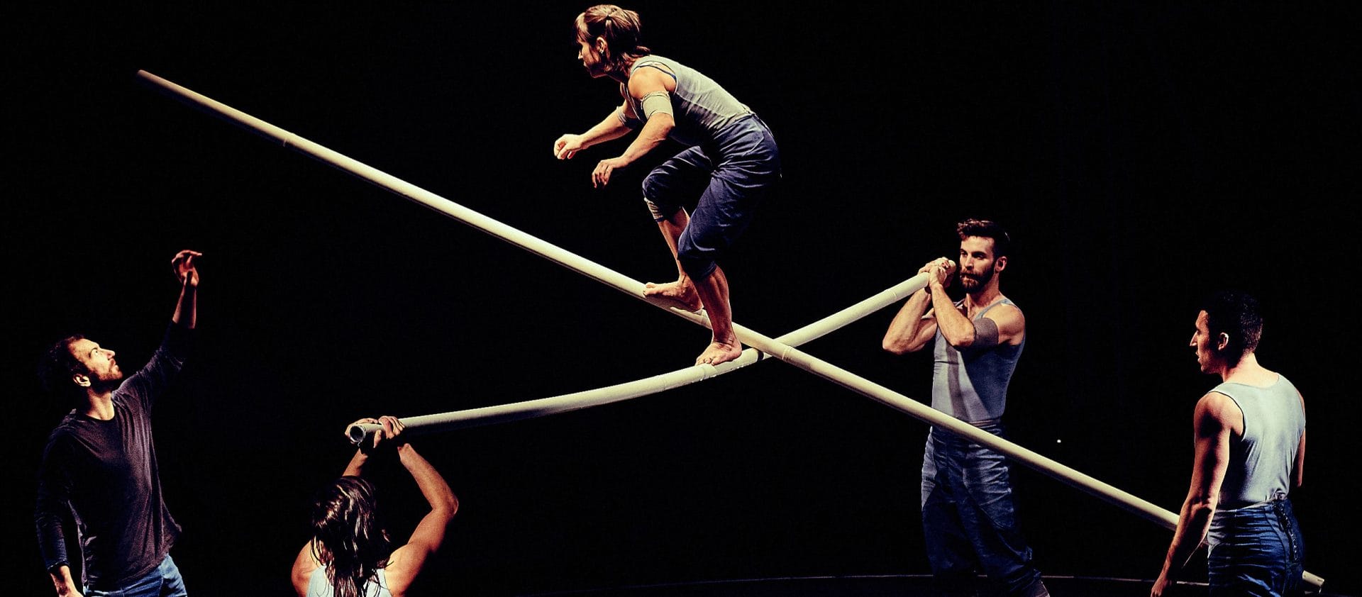three performers hold two long scaffold poles between them creating a cross, a fourth performer climbs on the poles while a fifth watches, they are on an indoor stage and all wear short blue jeans and pale blue vests