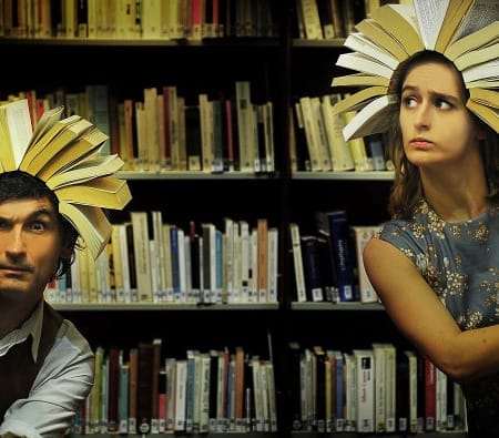 Two individuals stand between tall bookshelves in a dimly lit library, each wearing an open book as a headpiece, resembling sunbursts or crowns. One person, dressed in a vest and white shirt, peers curiously from behind a shelf, while the other in a vintage floral dress glances sideways with a concerned expression.