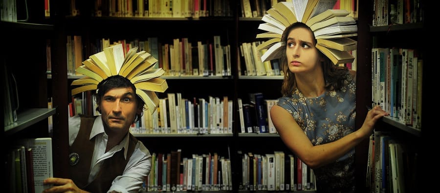 Two individuals stand between tall bookshelves in a dimly lit library, each wearing an open book as a headpiece, resembling sunbursts or crowns. One person, dressed in a vest and white shirt, peers curiously from behind a shelf, while the other in a vintage floral dress glances sideways with a concerned expression.