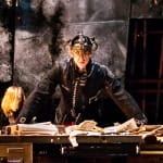 Performer behind a messy desk on an indoor stage, dressed in a dark uniform with an elaborate chandelier helmet looks at the camera, surrounded by other performers and softly lit by golden desk lamps James Thierrée Tabac Rouge