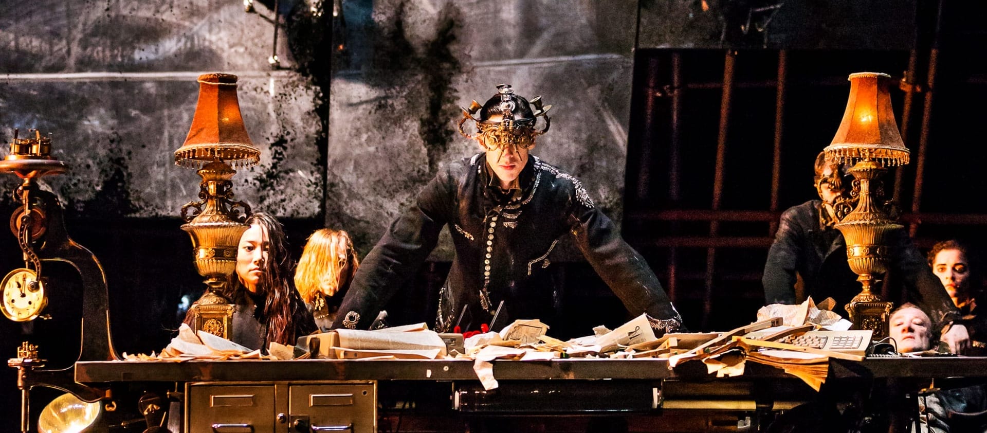 Performer behind a messy desk on an indoor stage, dressed in a dark uniform with an elaborate chandelier helmet looks at the camera, surrounded by other performers and softly lit by golden desk lamps James Thierrée Tabac Rouge