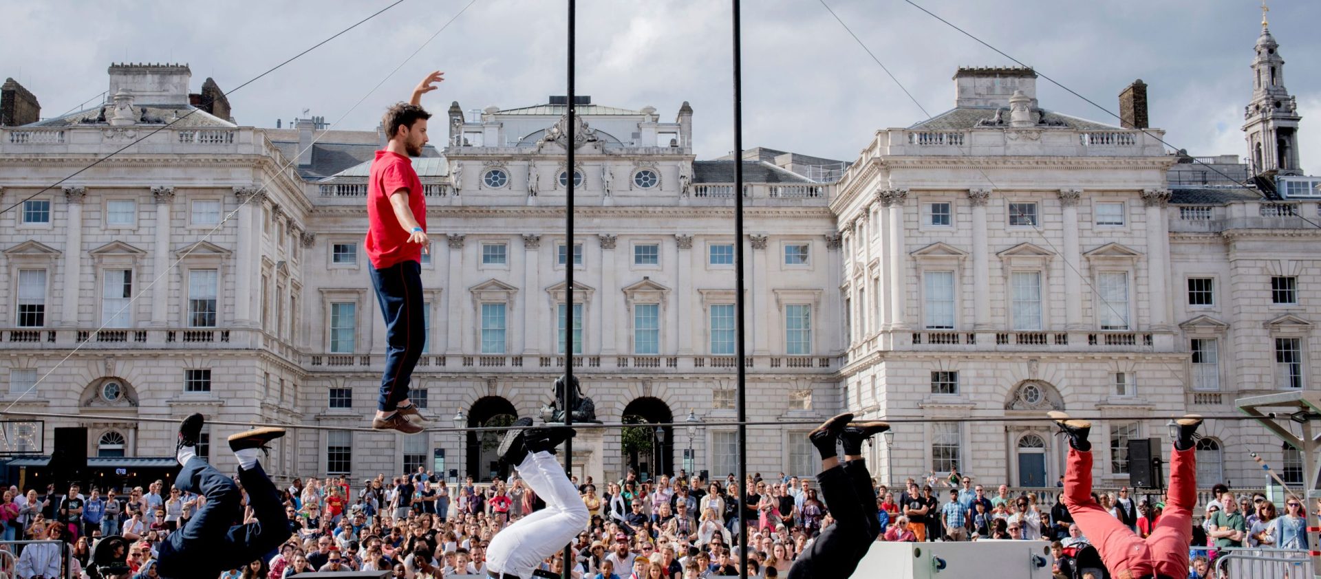 A tightrope walker and breakdancers perform before a crowd in front of a historic building.