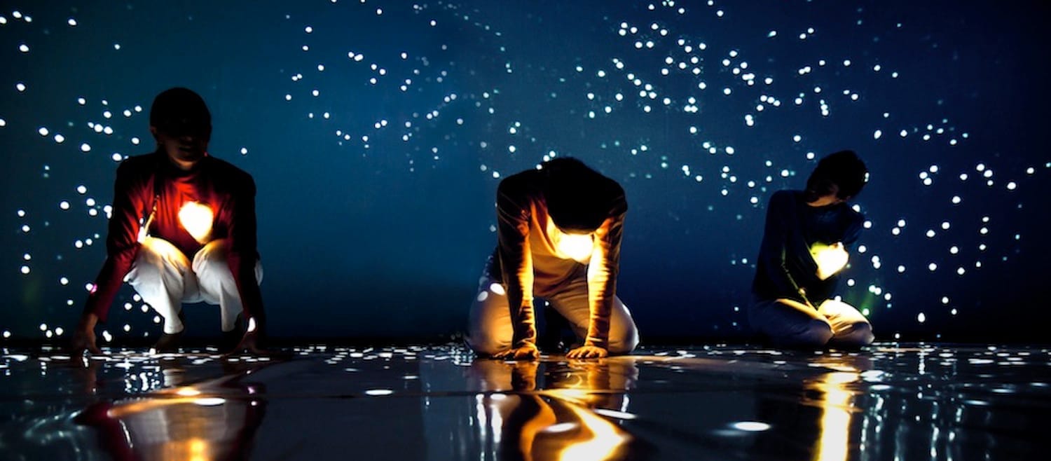 Three performers kneel on a dark reflective surface, surrounded by sparkling lights against a deep blue backdrop.
