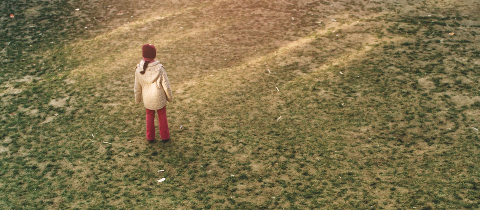 A person in a cream-coloured coat, red hat, and red trousers stands alone in a field of dull green grass.