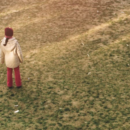 A person in a cream-coloured coat, red hat, and red trousers stands alone in a field of dull green grass.