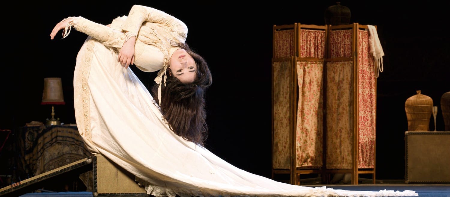 A performer in a flowing vintage cream-coloured gown bends gracefully backward, their hair cascading as ornate props stand against a dark stage background.  James Thierrée Veillée Abysses