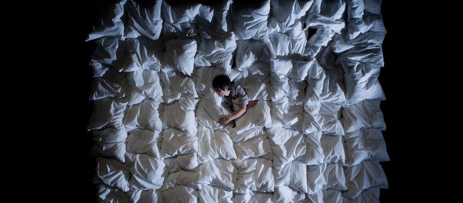 A person lies among a sea of white pillows, creating a soft, textured backdrop against a dark environment.