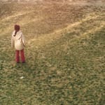 A person in a cream-coloured coat, red hat, and red trousers stands alone in a field of dull green grass.