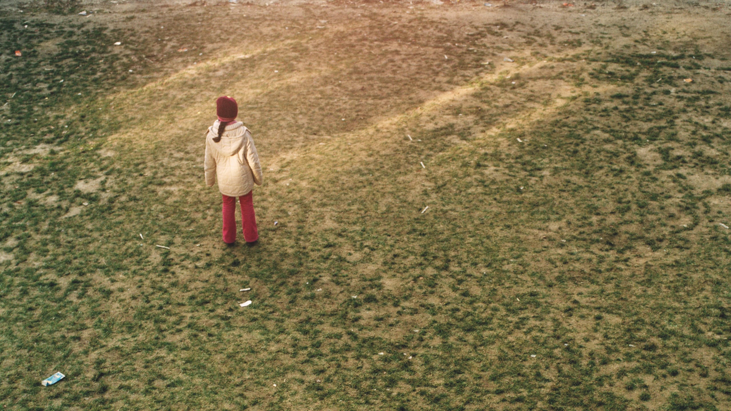 A person in a cream-coloured coat, red hat, and red trousers stands alone in a field of dull green grass.
