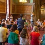 A lively gathering in an ornate hall, where participants sit in a circle, surrounding two presenters dressed in colourful outfits at the centre.