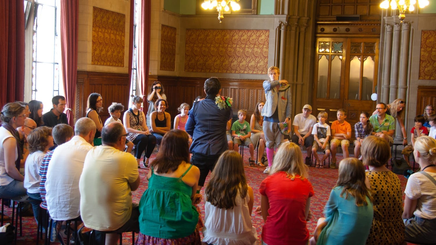 A lively gathering in an ornate hall, where participants sit in a circle, surrounding two presenters dressed in colourful outfits at the centre.