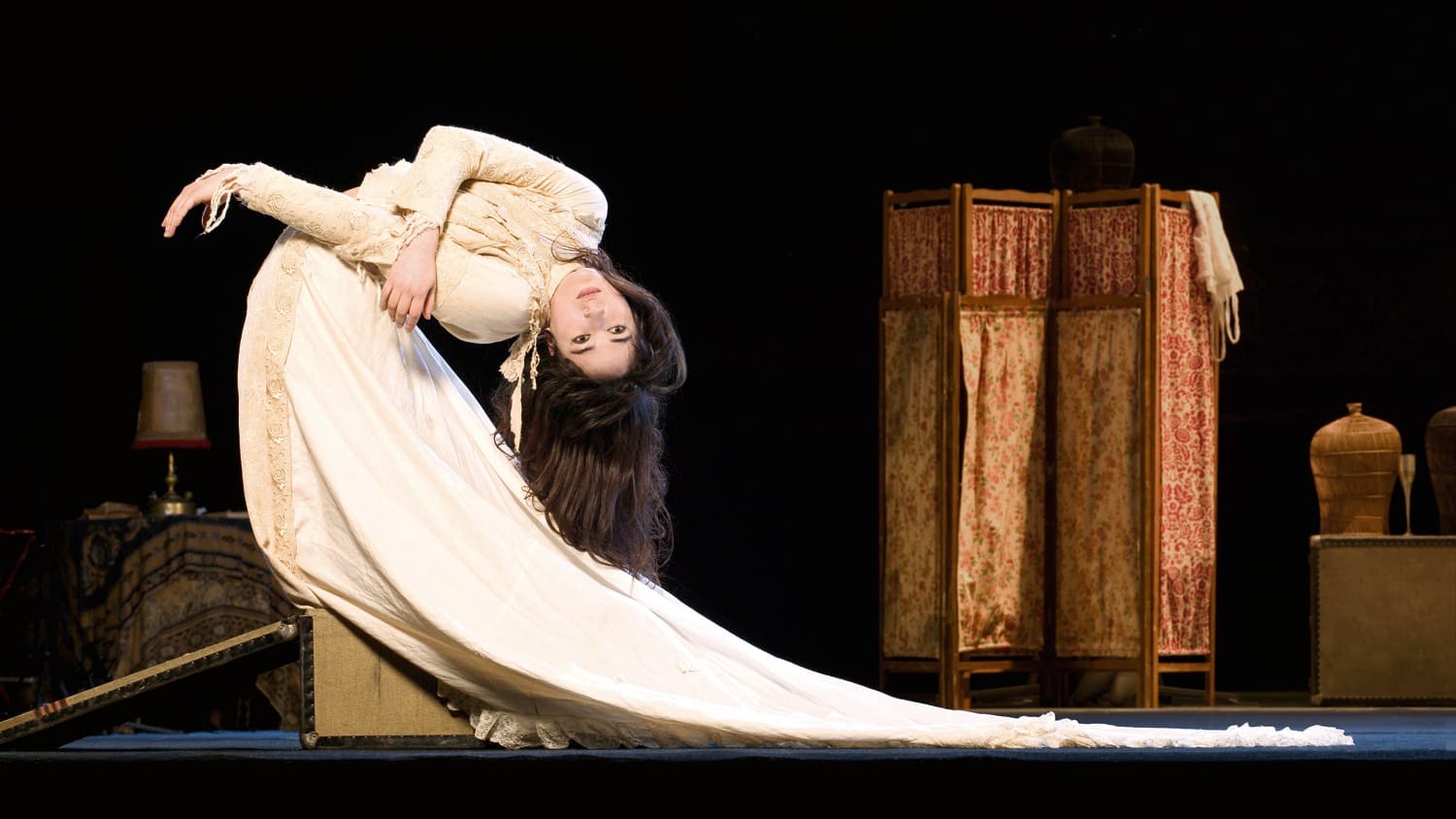 A performer in a flowing vintage cream-coloured gown bends gracefully backward, their hair cascading as ornate props stand against a dark stage background.