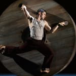 A person performs acrobatic moves on a large wooden wheel against a dark background, showcasing strength and agility.