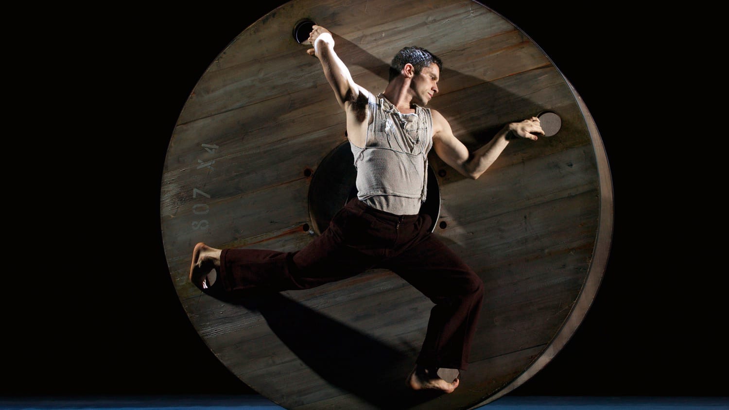 A person performs acrobatic moves on a large wooden wheel against a dark background, showcasing strength and agility.