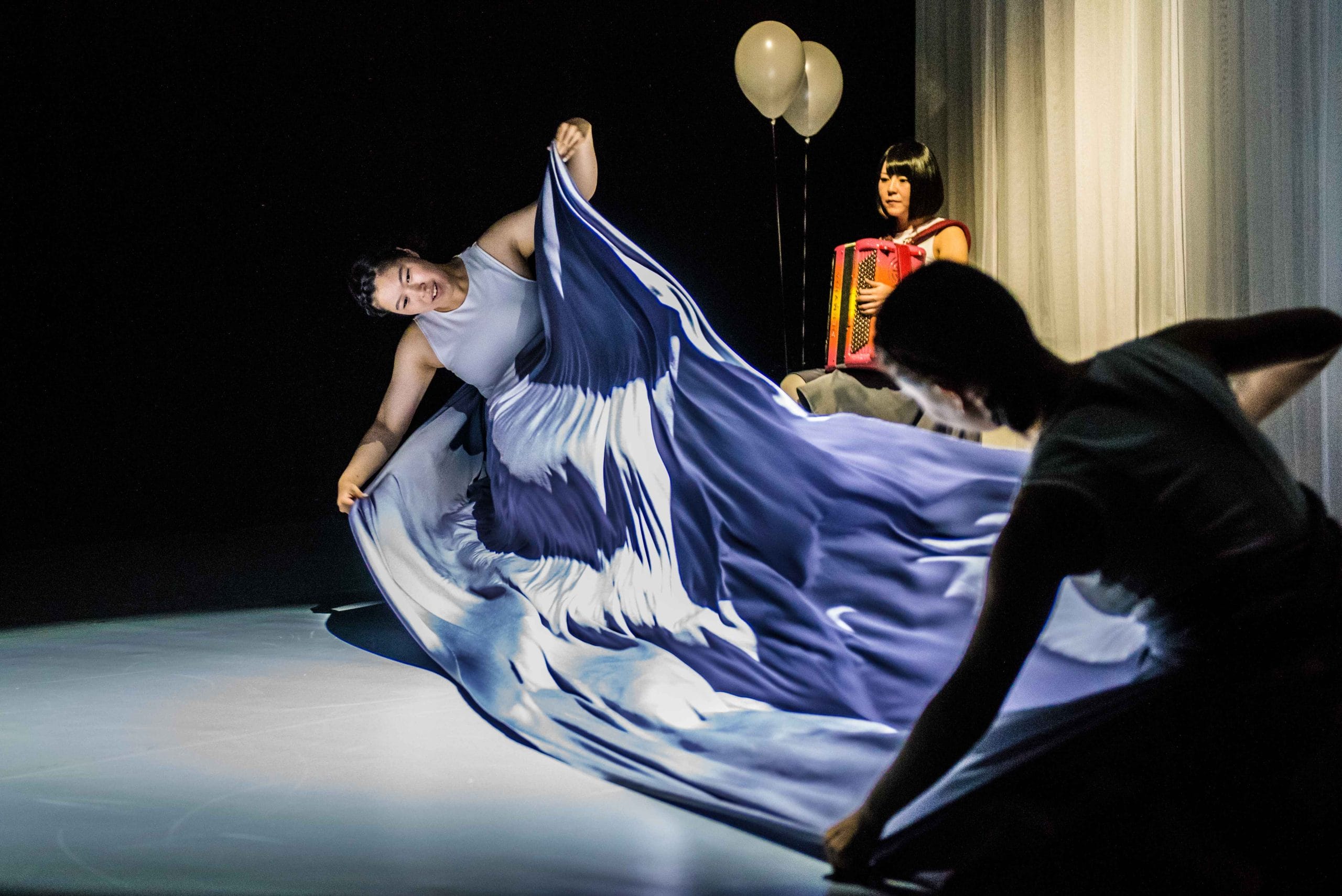 Three performers on stage, illuminated against a dark background. Two dancers in flowing costumes manipulate a large piece of fabric with a blue and white wave-like pattern, creating a dynamic visual effect. In the background, a musician in a sleeveless top plays a red accordion, seated near sheer white curtains and two white balloons floating above.