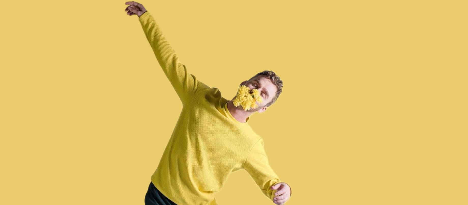 a person with short brown hair wearing a yellow sweater, against a yellow background has a yellow dandelion in their mouth and holds both arms out the side movement director Luke Brown