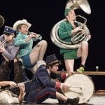 A lively band of seven musicians in colourful, mismatched outfits playing various instruments on stage, showcasing energetic movement and creativity.