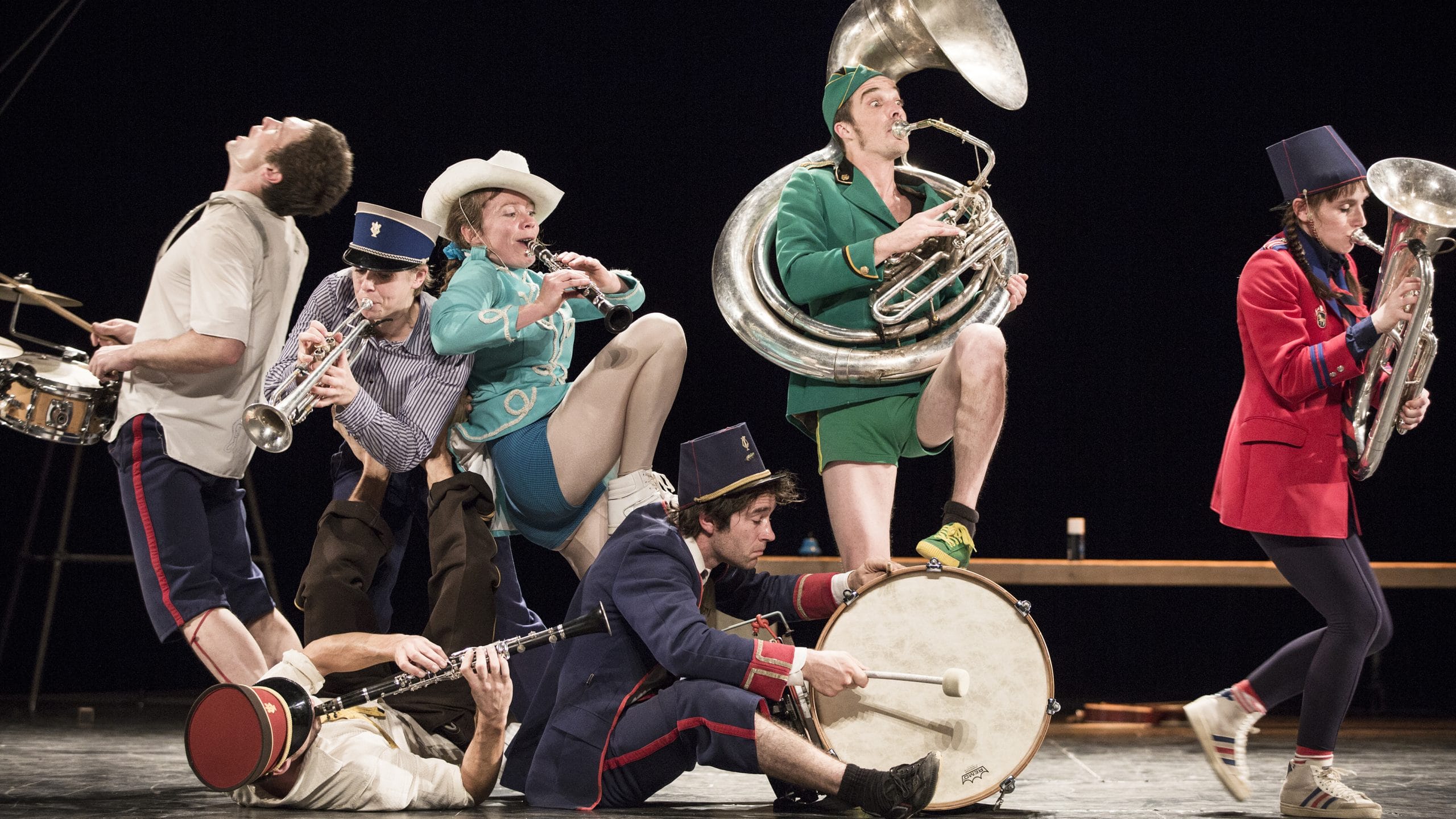 A lively band of seven musicians in colourful, mismatched outfits playing various instruments on stage, showcasing energetic movement and creativity.