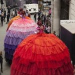 three performers on high stilts wearing elaborately coloured, many layered, petticoats walk through a crowded narrow walkway with grand stone building either side