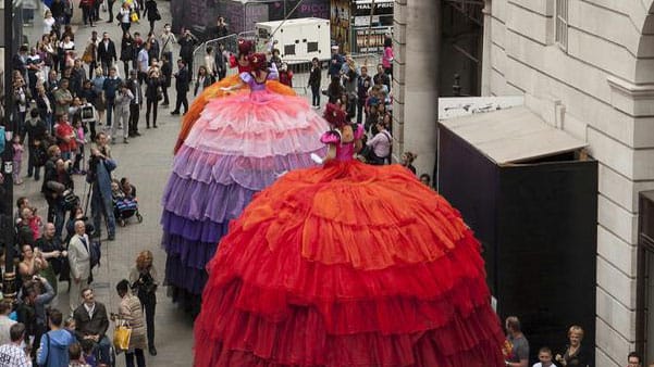 three performers on high stilts wearing elaborately coloured, many layered, petticoats walk through a crowded narrow walkway with grand stone building either side