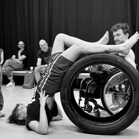 black and white image of an indoor rehearsal space with three performers moving their bodies over large car tyres, with three onlookers