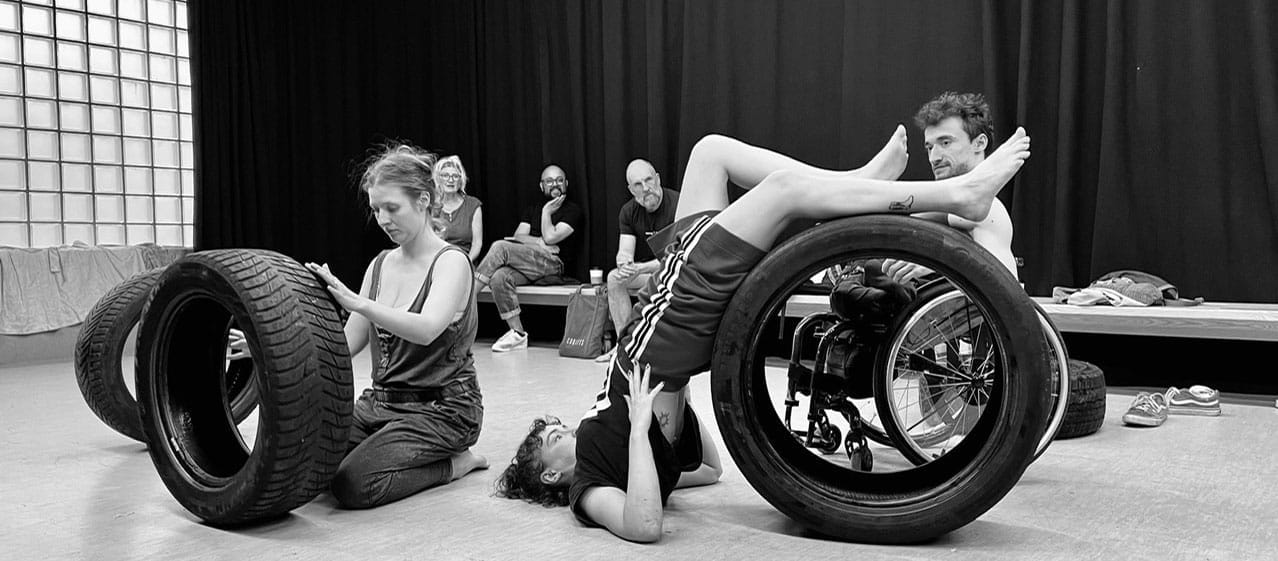 black and white image of an indoor rehearsal space with three performers moving their bodies over large car tyres, with three onlookers