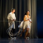 performer with long loose dark hair, wearing a short floaty white sleeveless dress, runs away as two figures in lab coats hold onto ropes, creating a dramatic performance scene.