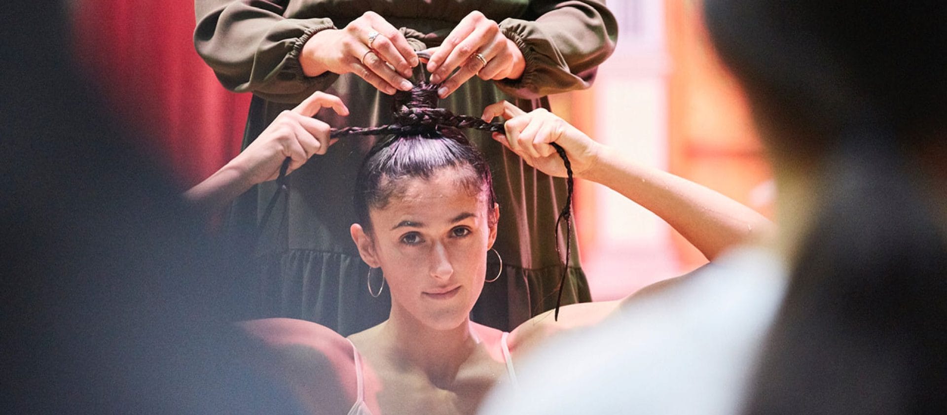 A hair-hanging performer ties their dark, plaited hair in a top-know, they wear a white scrappy vest top and a second person in a green dress assists.