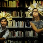 Two individuals stand between tall bookshelves in a dimly lit library, each wearing an open book as a headpiece, resembling sunbursts or crowns. One person, dressed in a vest and white shirt, peers curiously from behind a shelf, while the other in a vintage floral dress glances sideways with a concerned expression.