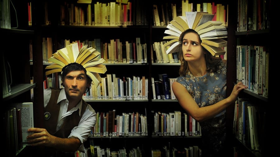 Two individuals stand between tall bookshelves in a dimly lit library, each wearing an open book as a headpiece, resembling sunbursts or crowns. One person, dressed in a vest and white shirt, peers curiously from behind a shelf, while the other in a vintage floral dress glances sideways with a concerned expression.