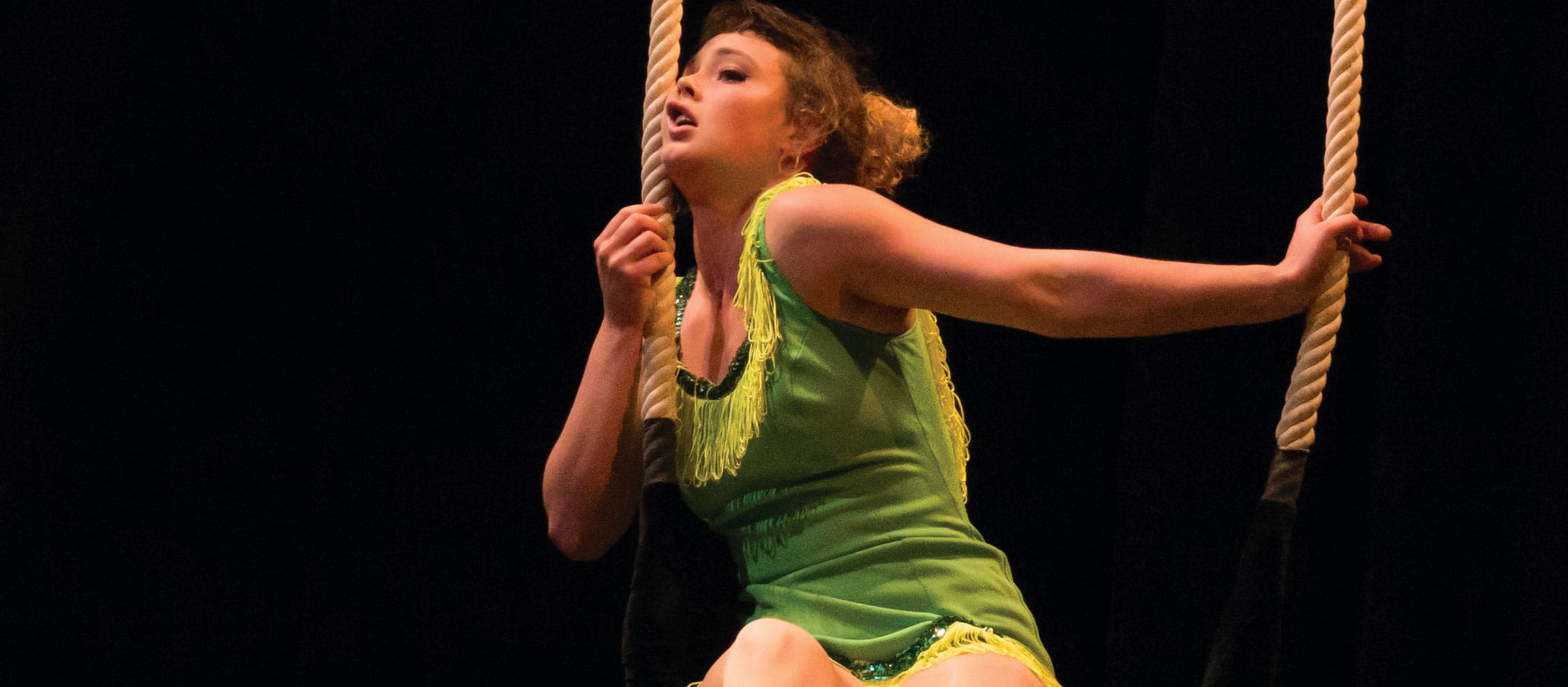 Aerial performer on an indoor stage on a trapeze wearing a green costume with hassled paler green trim