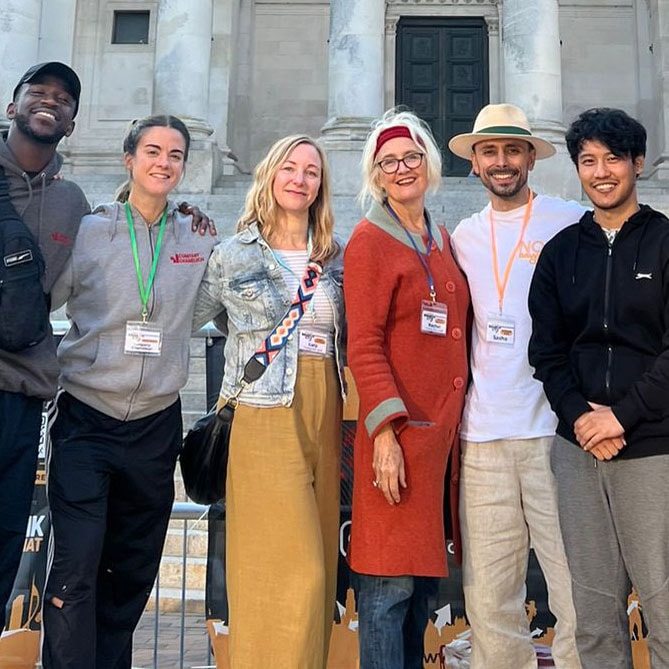 six people of differing ethnicities wearing colourful clothing and matching lanyards stand in front of a grand grey stone building smiling at the camera