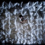 A person lies among a sea of white pillows, creating a soft, textured backdrop against a dark environment.