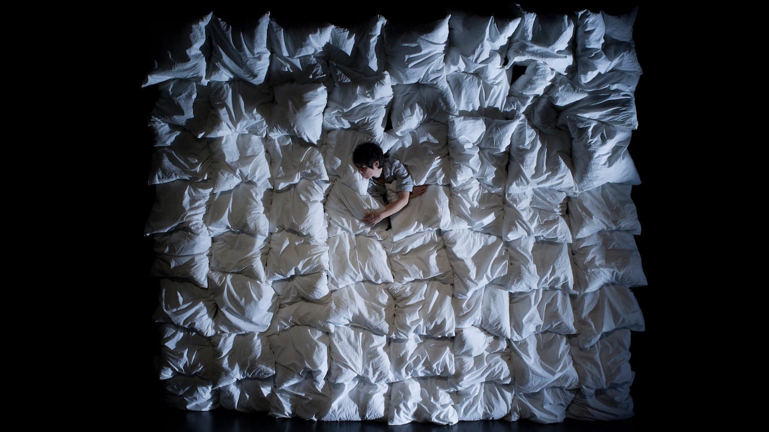 A person lies among a sea of white pillows, creating a soft, textured backdrop against a dark environment.