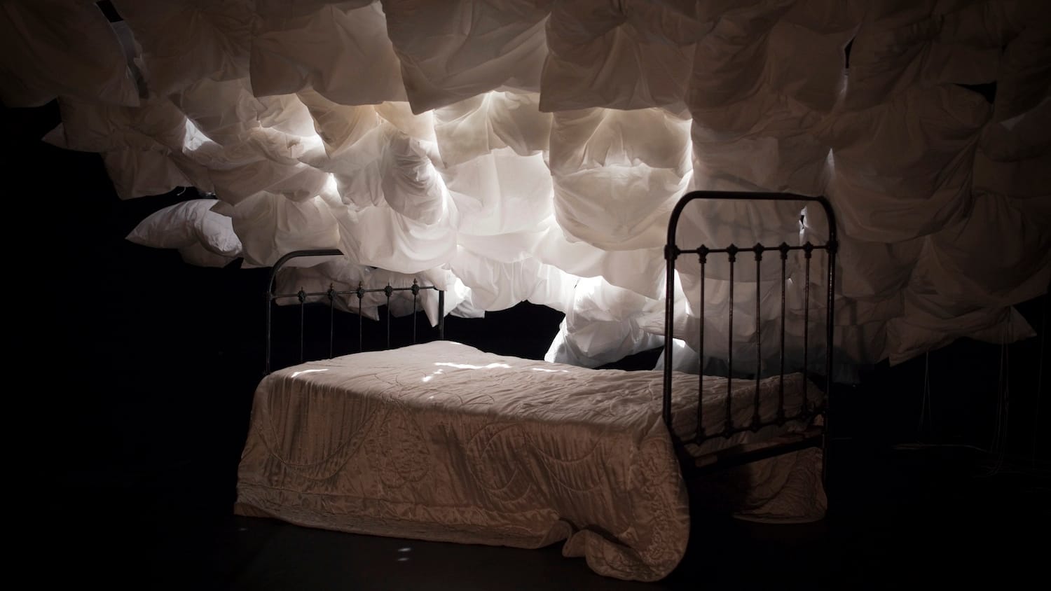 A vintage bed with black frame and white blanket stands beneath a cloud-like arrangement of white pillows, illuminated by soft white light in a dark space.