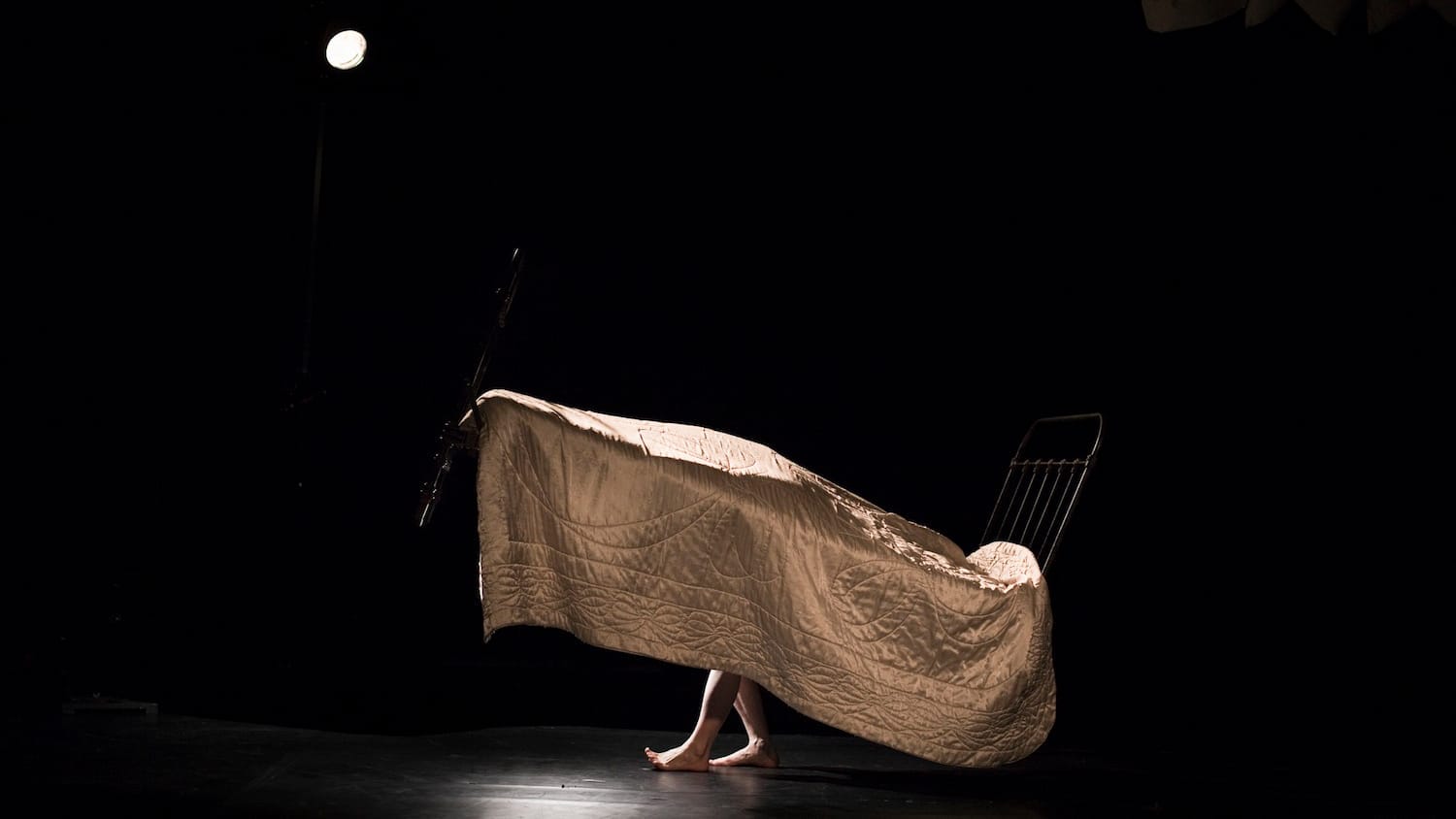 A dimly lit stage features a vintage black bed covered with a quilt and a pair of bare feet peeking out, suggesting a hidden presence beneath the fabric.