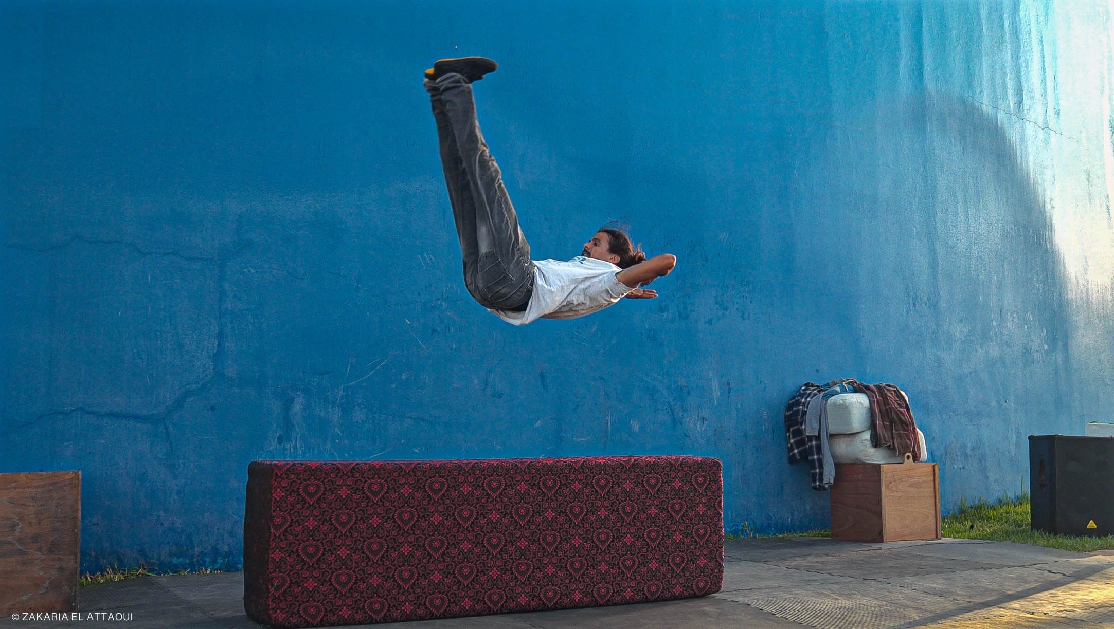an acrobat dressed in white t-shirt and blue jeans with long dark hair and dark facial hair is bouncing on a piece of flat furniture set up outdoors against a backdrop of a blue wall, their feet are in the air and hands behind their head