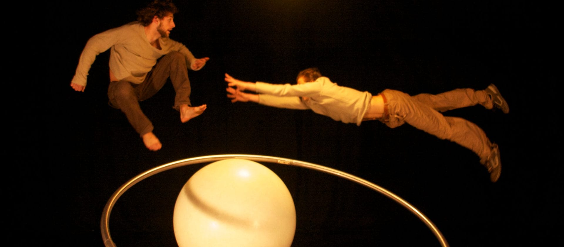 two acrobats both with brown hair and beards, wearing brown trousers and grey sweatshirts perform over a gym ball and with a cyr wheel in an indoor stage setting with yellow lighting