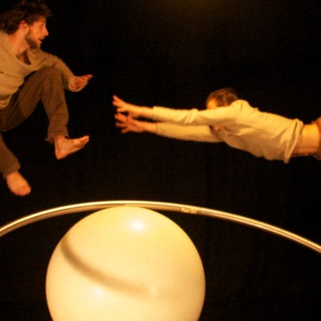 two acrobats both with brown hair and beards, wearing brown trousers and grey sweatshirts perform over a gym ball and with a cyr wheel in an indoor stage setting with yellow lighting
