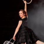 A performer in a black dress hanging gracefully from an aerial hoop above an iron board, set against a misty background.