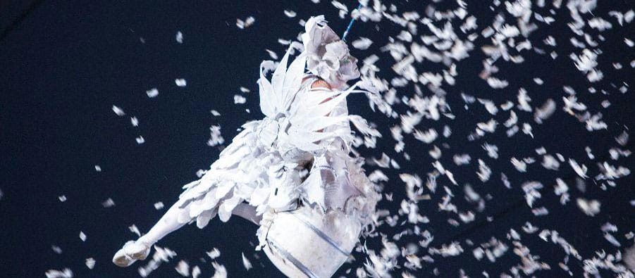 an aerial performer dressed as an angel in a white outfit with wings, is suspended on a highwire, releasing handfuls of feathers which erupt against a dark sky Piccadilly Circus Circus