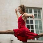 a tightrope walker in a red dress performs the splits on an outdoor highwire against a backdrop of a grand building