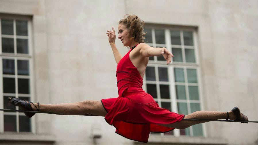 a tightrope walker in a red dress performs the splits on an outdoor highwire against a backdrop of a grand building