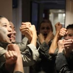 Six performers in a backstage corridor, all with roughly applied white face makeup, dressed in a dark trousers and grey shirts move as one with their forearms crossed in front of them James Thierrée Tabac Rouge