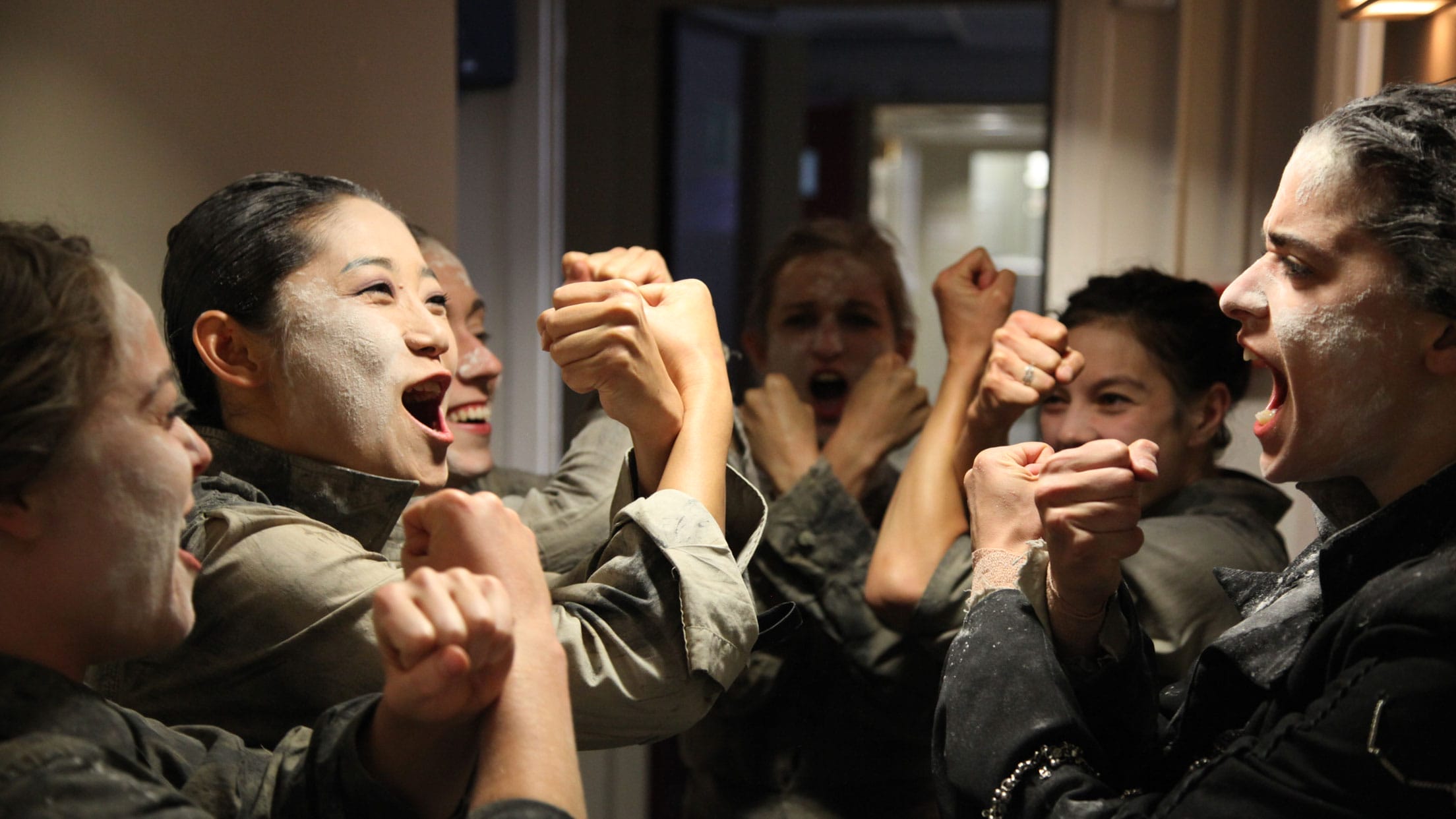 Six performers in a backstage corridor, all with roughly applied white face makeup, dressed in a dark trousers and grey shirts move as one with their forearms crossed in front of them James Thierrée Tabac Rouge