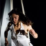 performer with long loose dark hair, wears a short floaty white sleeveless dress in an indoor darkened stage, they are suspended mid-air by ropes